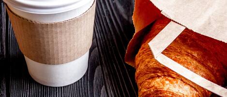 Una taza de café con tapa y una bolsa de papel que contiene un croissant sobre una superficie de madera.