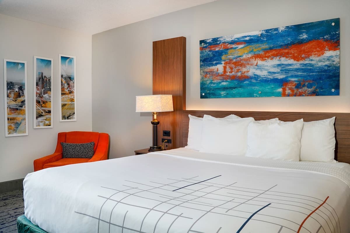 A hotel room with a large bed, a nightstand with a lamp, a chair, and three framed cityscape pictures on the wall.