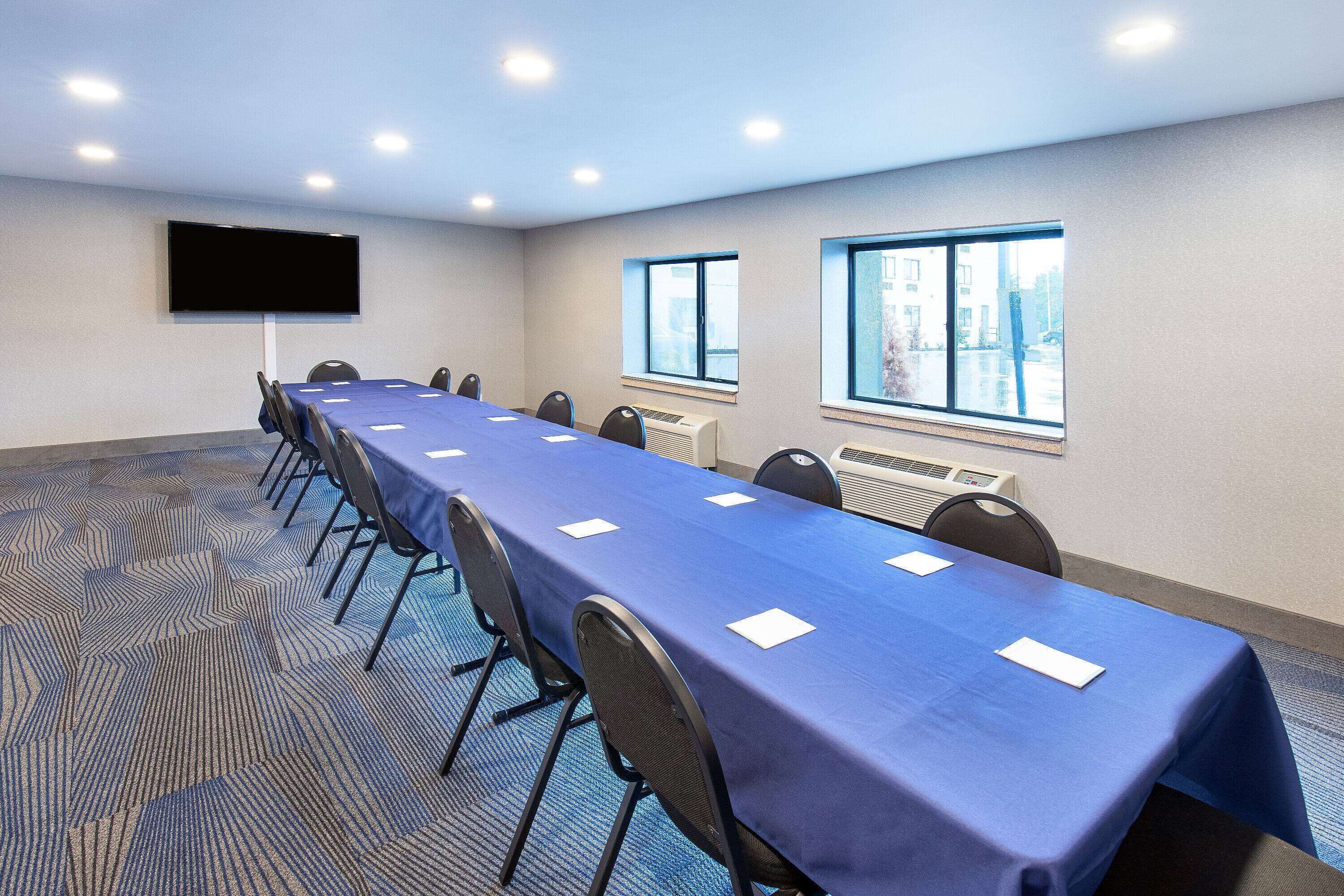 Un salón de conferencias con una larga mesa, sillas, un televisor de pantalla plana, ventanas y un equipo de aire acondicionado.