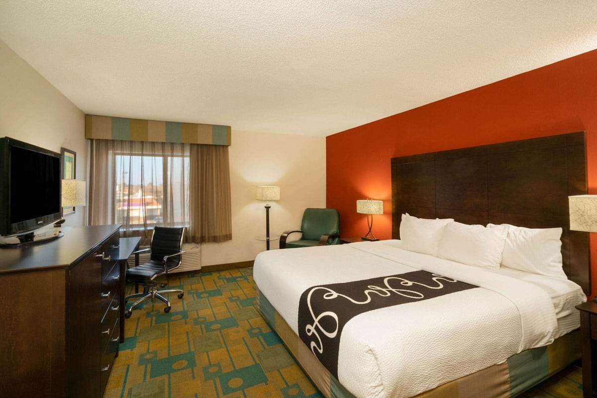 A hotel room with a bed, a desk with a TV, a chair, a lamp, and a view of the city.