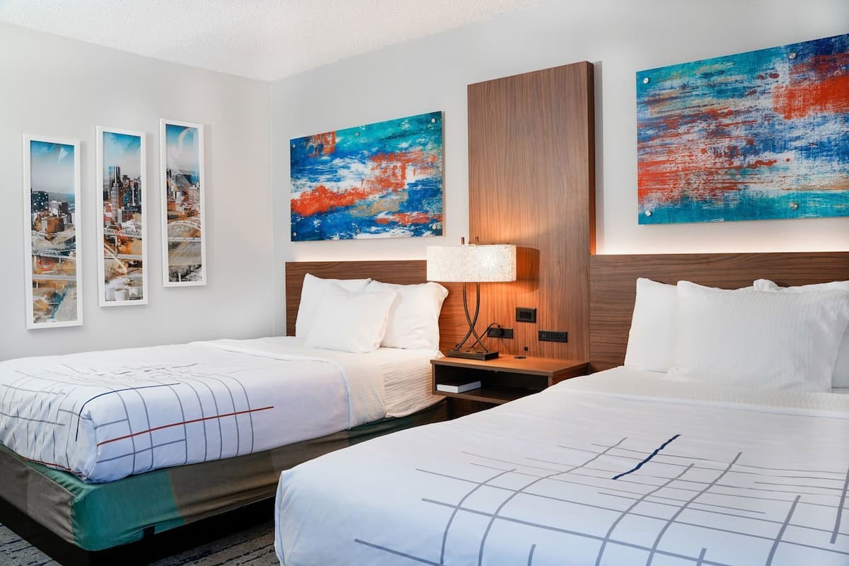 A hotel room with two beds, a nightstand, a lamp, and three framed cityscape pictures on the wall.