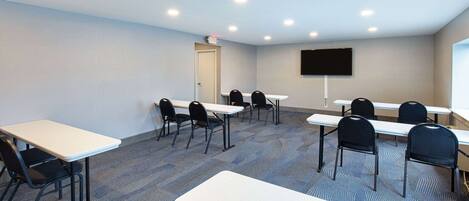 A conference room with multiple tables and chairs, a flat-screen TV mounted on the wall, and a carpeted floor.