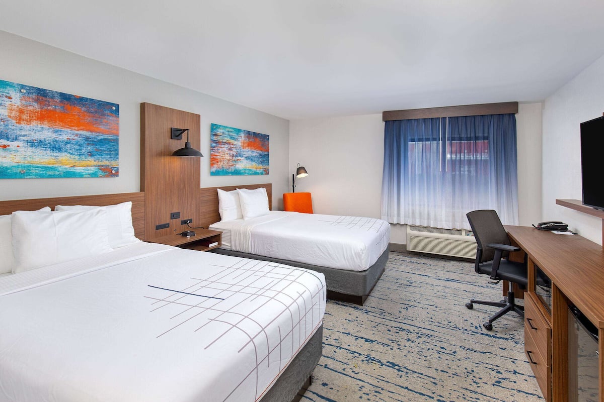 A hotel room with two beds, a desk, a chair, a TV, and artwork on the walls.