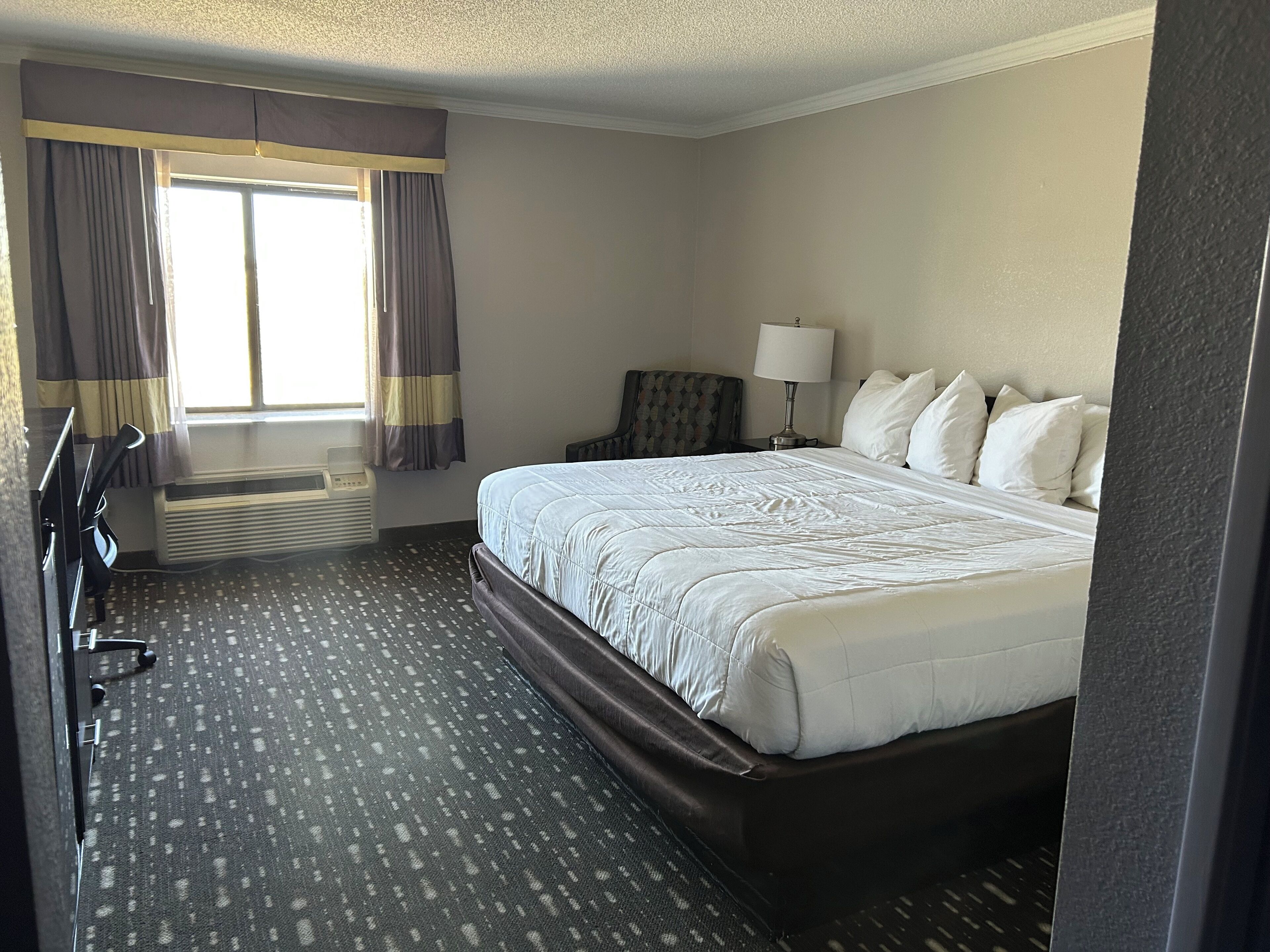 Una camera d'albergo con un letto, una scrivania, una sedia, una lampada e una finestra con tende.
