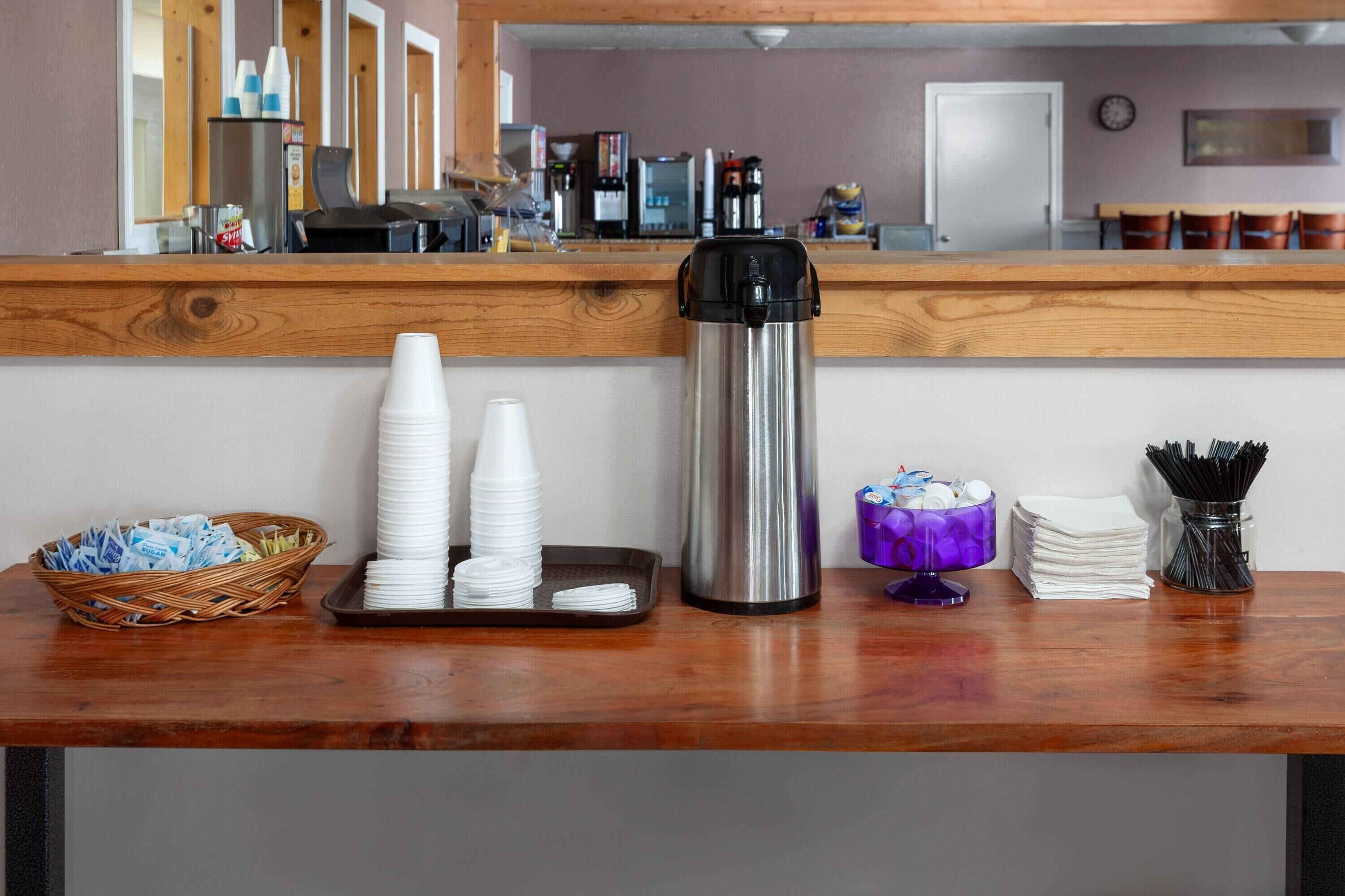 A reception area with a wooden counter, stacks of white cups, a stainless steel coffee pot, a wicker basket, and a purple container with utensils.