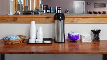 Uma área de recepção com balcão de madeira, pilhas de copos brancos, uma cafeteira de aço inoxidável, uma cesta de vime e um recipiente roxo com utensílios.