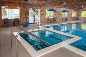 An indoor swimming pool with a brick wall, blue chairs, and umbrellas. - Baymont by Wyndham Manistee (Manistee)