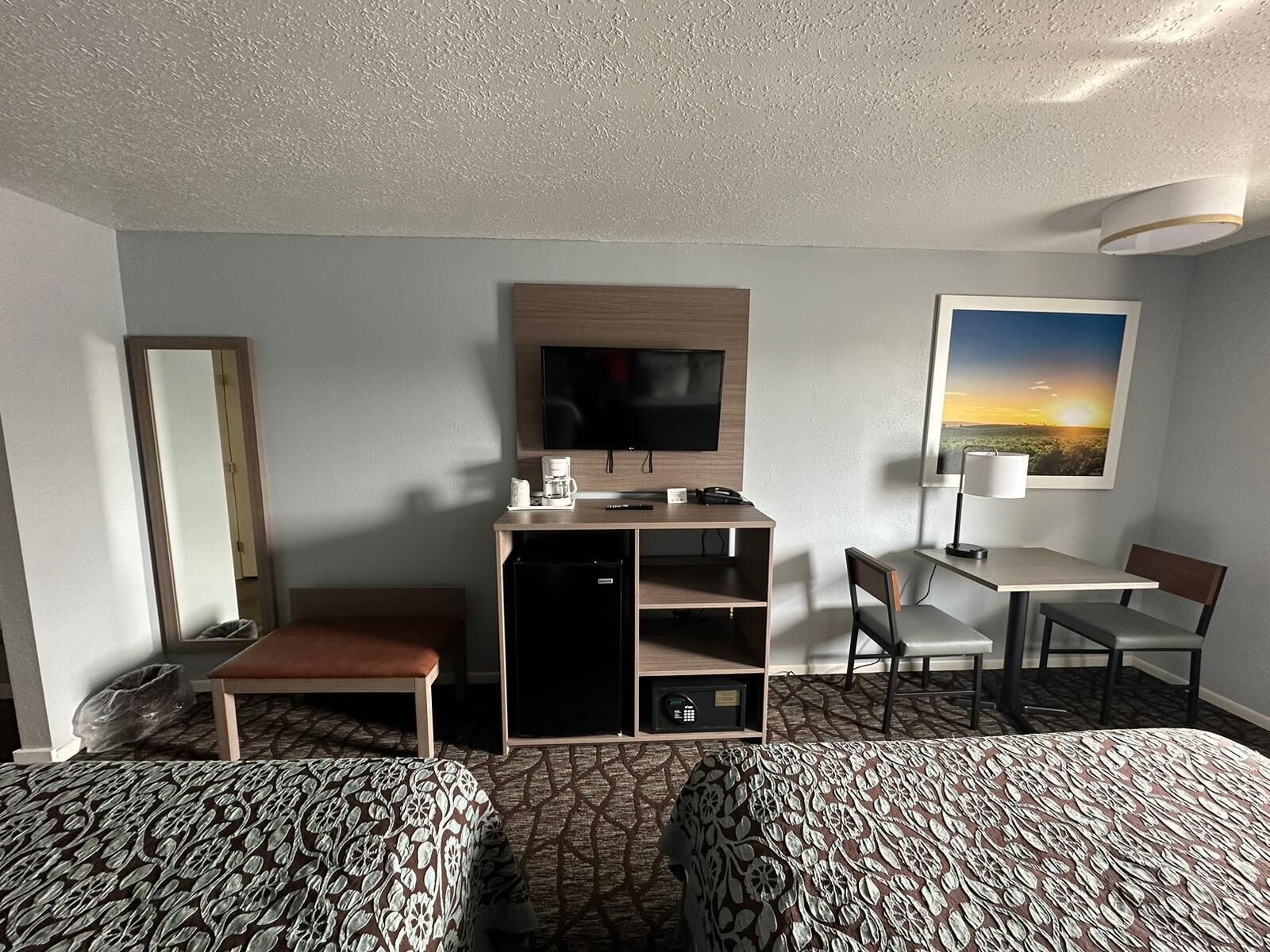 Quarto de hotel com duas camas, uma mesa de trabalho com uma televisão, uma cadeira, uma cafeteira e um quadro na parede.
