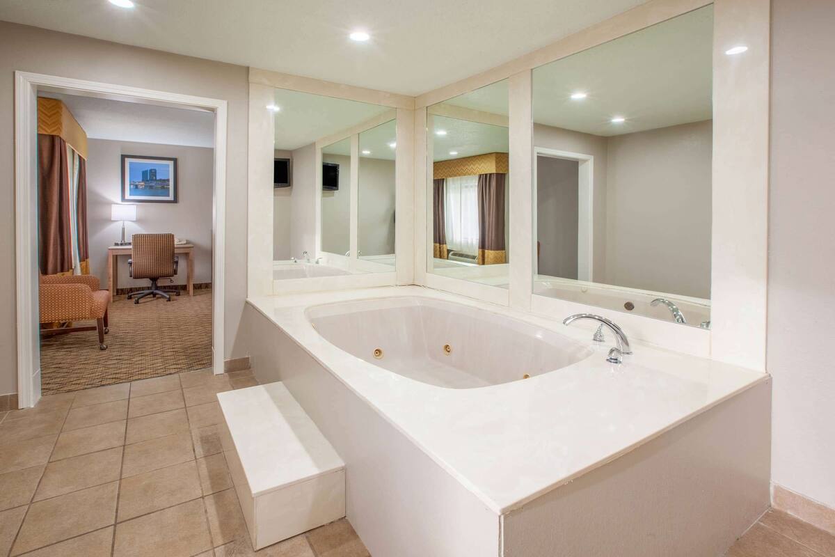 A modern bathroom with a large bathtub, a sink, and a mirror. There is a chair and a desk in the adjacent room.