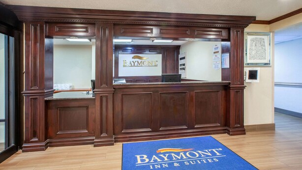 A reception desk with a 'Baymont Inn & Suites' sign, a blue mat at the entrance, and framed pictures on the wall. - Baymont by Wyndham Grand Rapids Airport (Grand Rapids)