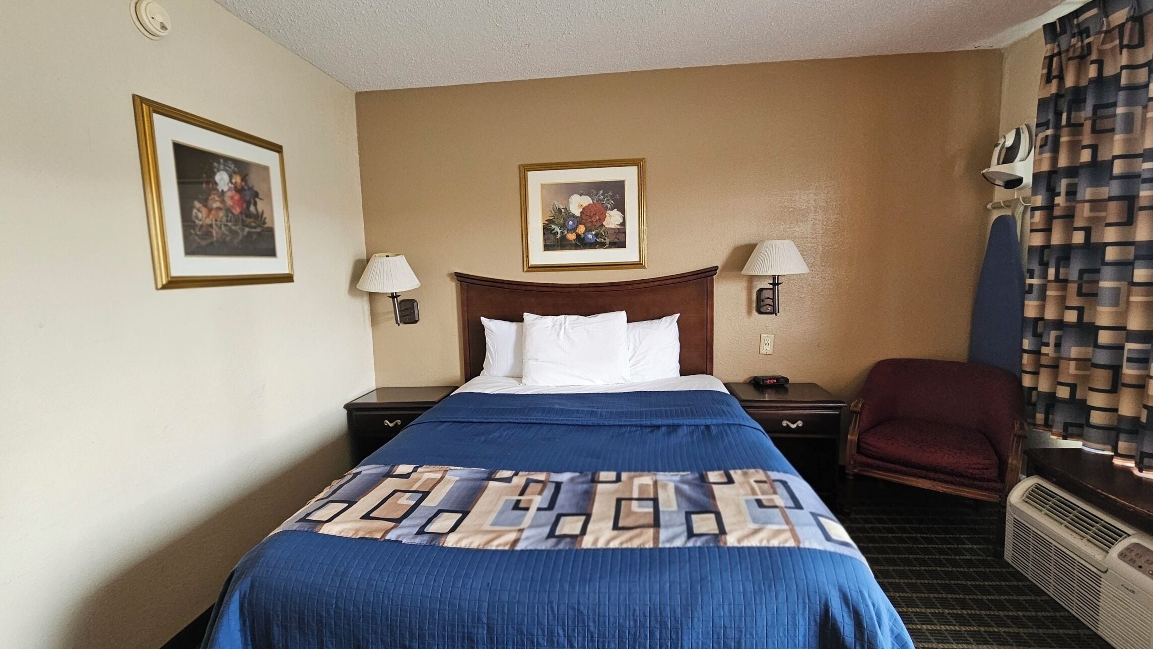 A hotel room with a bed, two bedside tables, a chair, a heater, and two wall-mounted lamps.