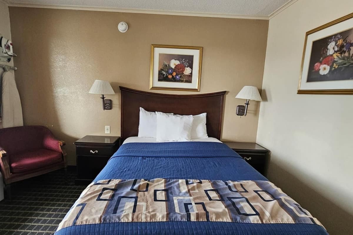 Une chambre d’hôtel avec un lit, deux lampes de chevet, une chaise, un radiateur et deux tableaux représentant des motifs floraux, accrochés au mur.