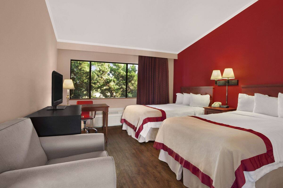 A hotel room with two beds, a desk with a computer, a chair, and a window with a view of trees.