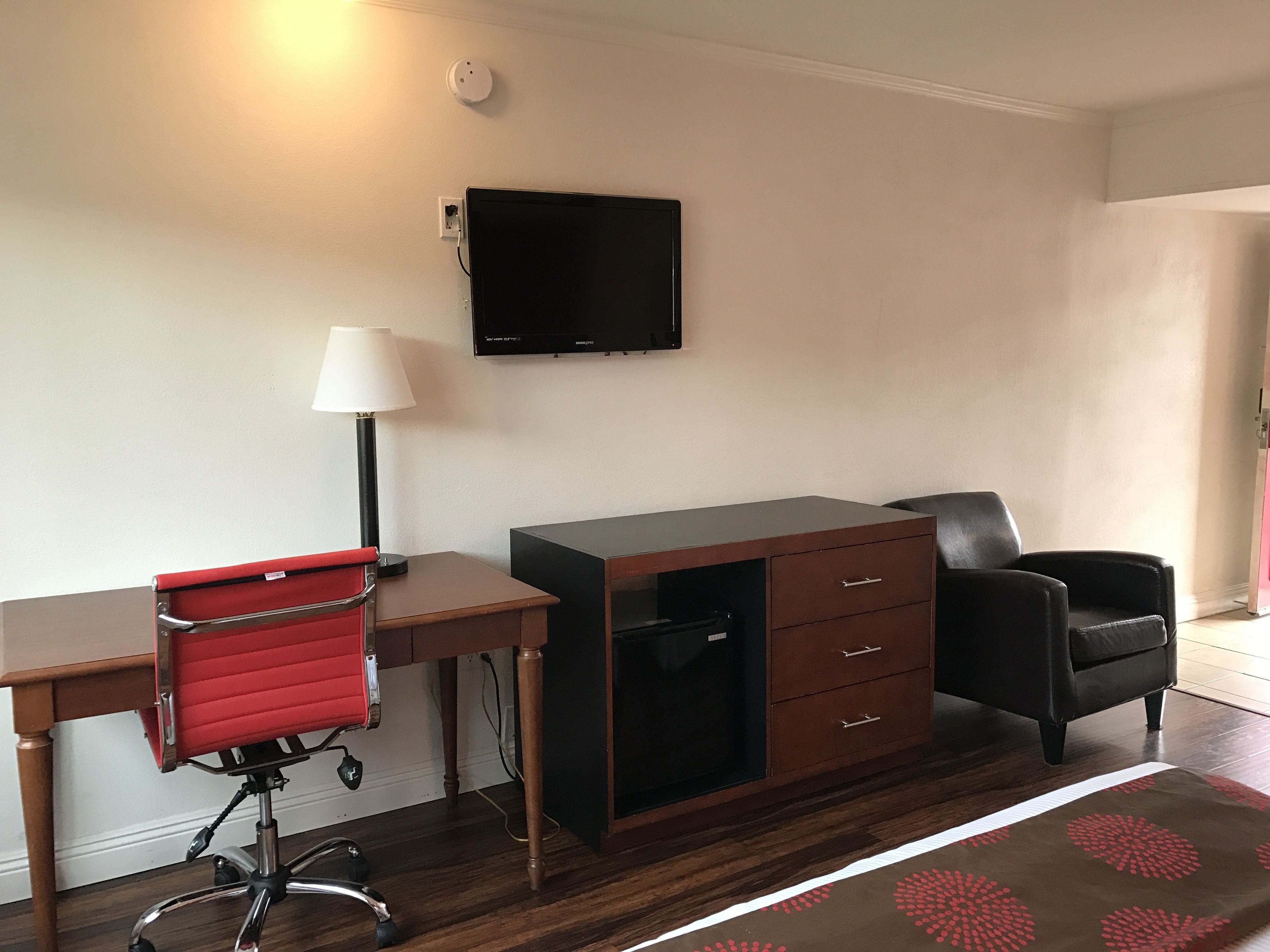 A hotel room with a wooden desk, a red office chair, a black leather chair, a TV mounted on the wall, and a patterned rug.