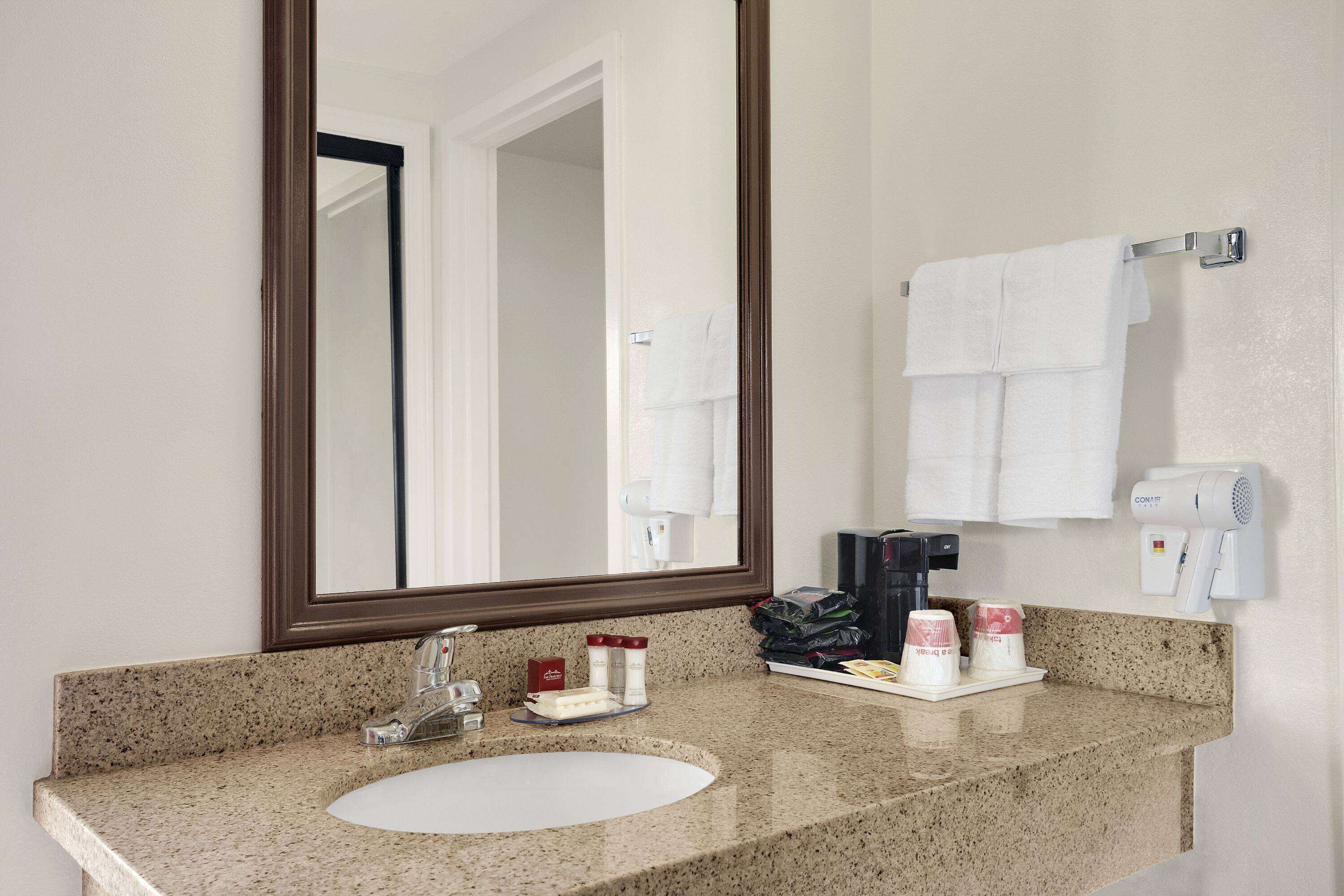 Baño de hotel con lavamanos de encimera de granito, un espejo grande y un perchero con varias toallas blancas.