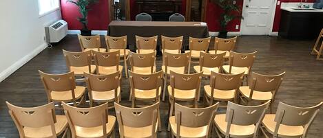 A conference room with a long table and multiple chairs arranged around it. There is a television on a stand in the background, and two potted plants are placed near the entrance.