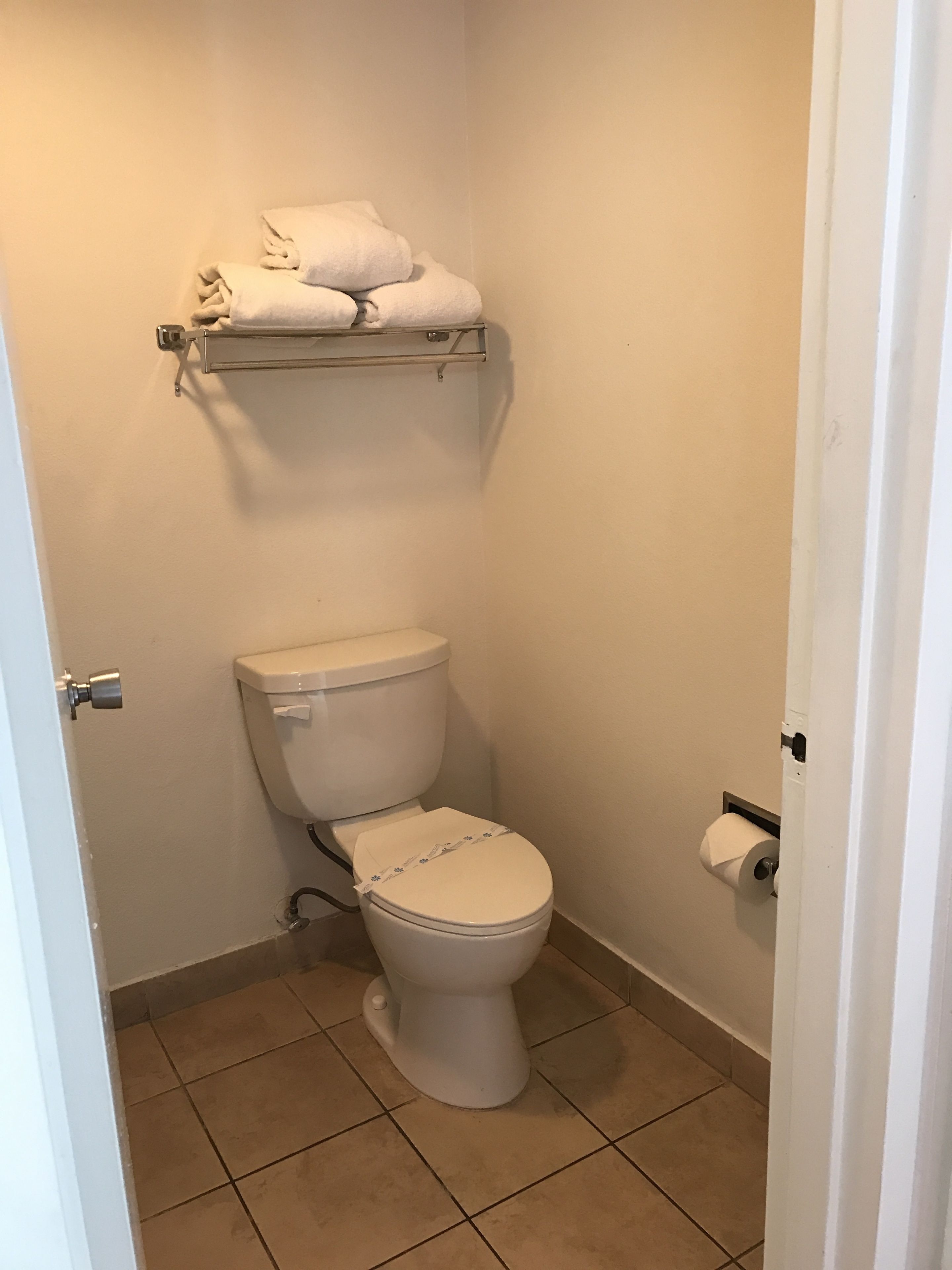 Un baño pequeño y limpio, con inodoro, un estante sobre el inodoro con algunas toallas y un dispensador de papel higiénico en la pared.