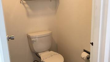 Un baño pequeño y limpio, con inodoro, un estante sobre el inodoro con algunas toallas y un dispensador de papel higiénico en la pared.