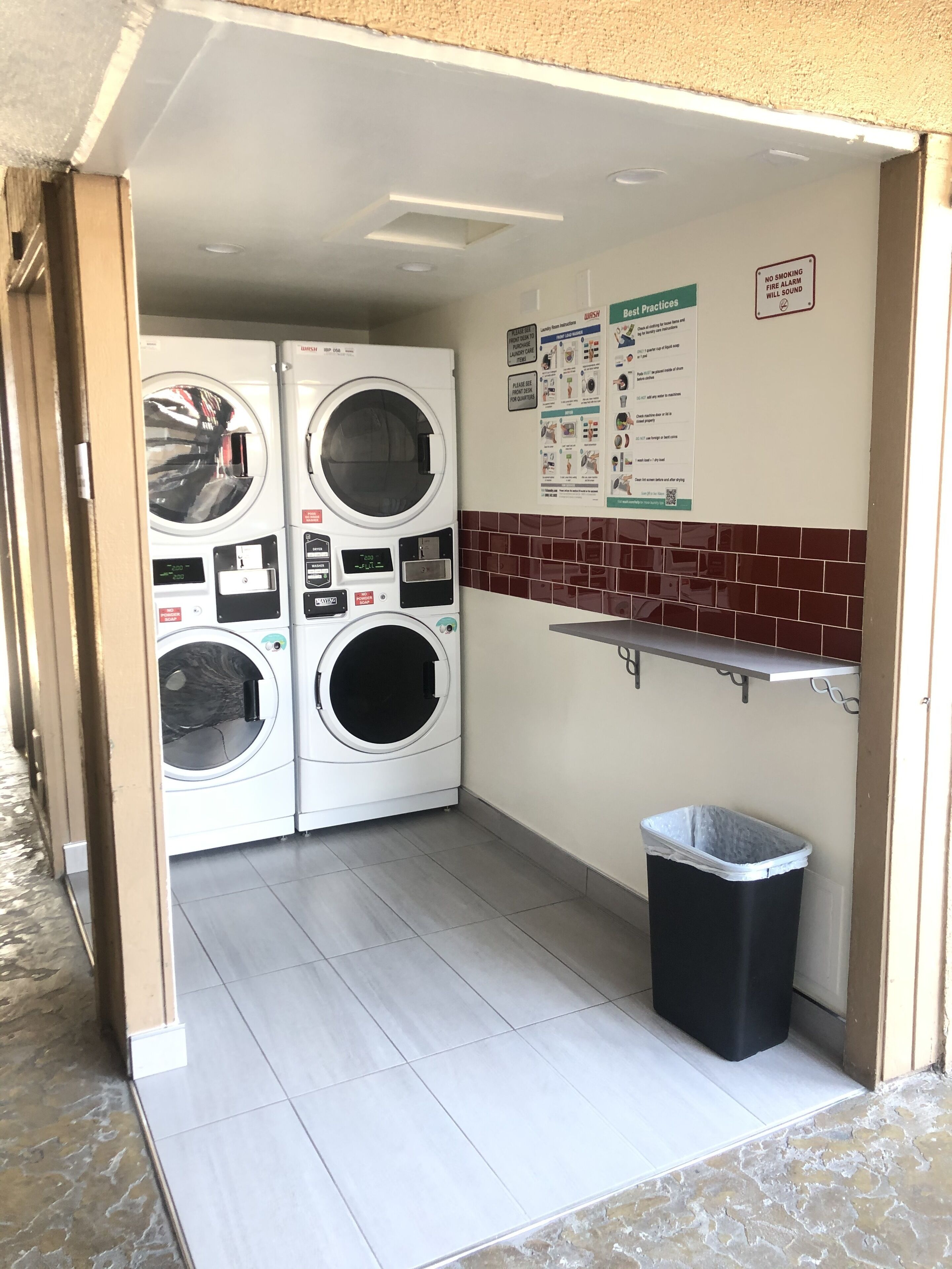 A laundry room with two front-loading washing machines, a trash bin, and a bulletin board with safety and laundry instructions.