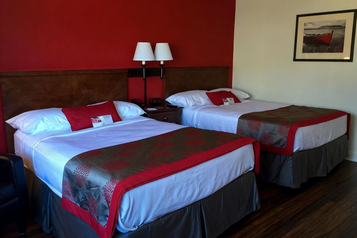 A hotel room with two beds, a red accent wall, a framed picture, and a nightstand with lamps.