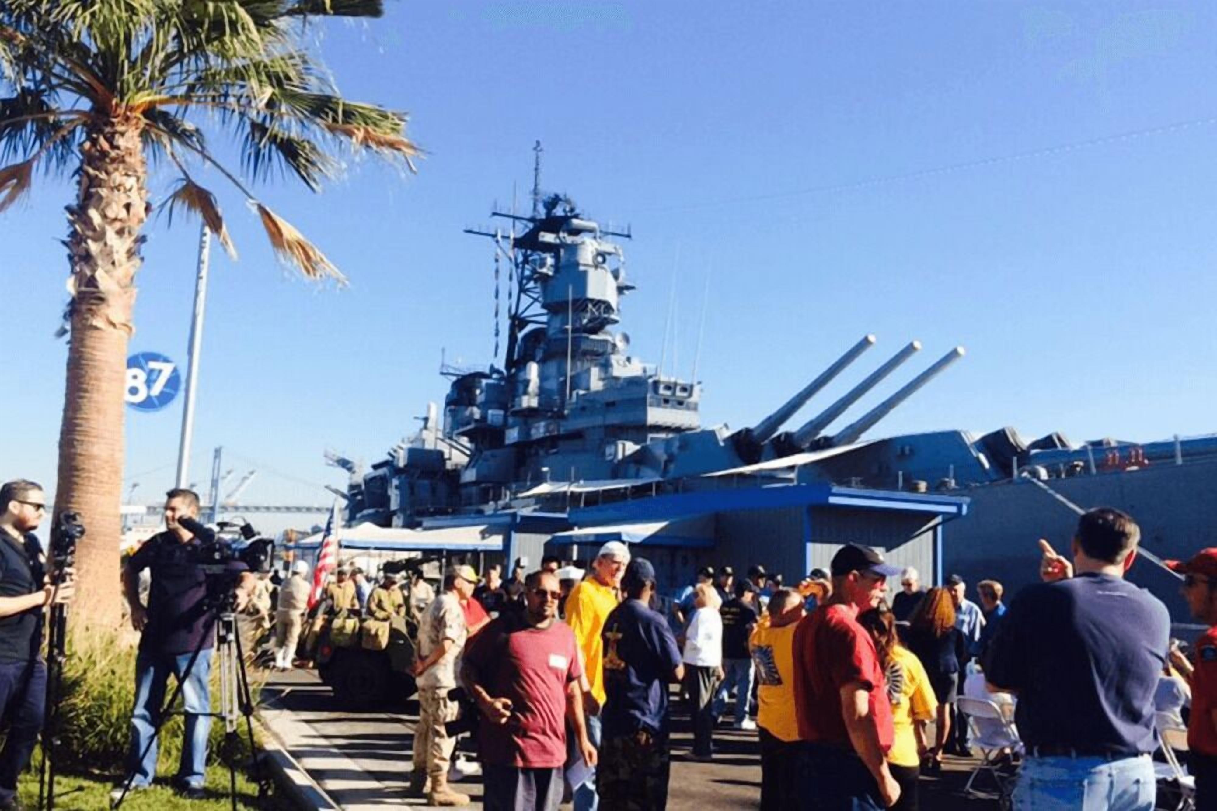 A large naval ship docked at a port with a crowd of people around it.