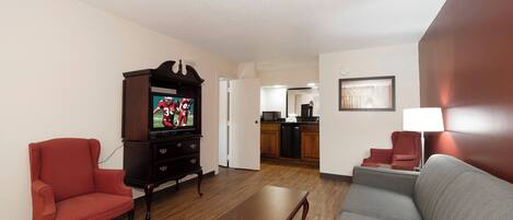 A living room with a television, a coffee table, two armchairs, and a kitchen area in the background.
