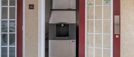 A compact ice and vending machine unit with a metal exterior, located in a doorway with a red door and a white-framed window on the left.