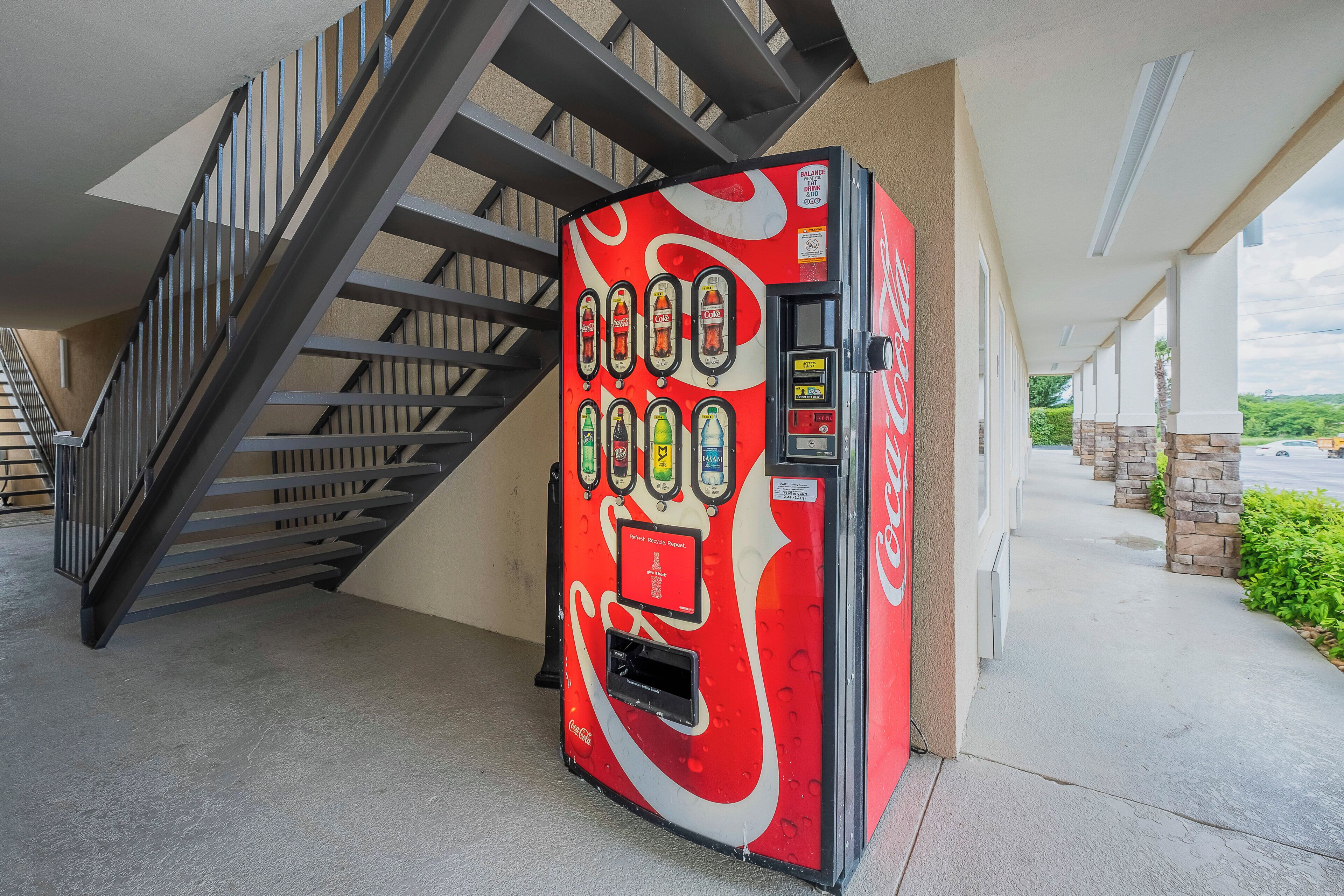 A Coca-Cola branded vending machine with a red and white design, located next to a staircase and a building exterior.