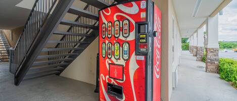A Coca-Cola branded vending machine with a red and white design, located next to a staircase and a building exterior.