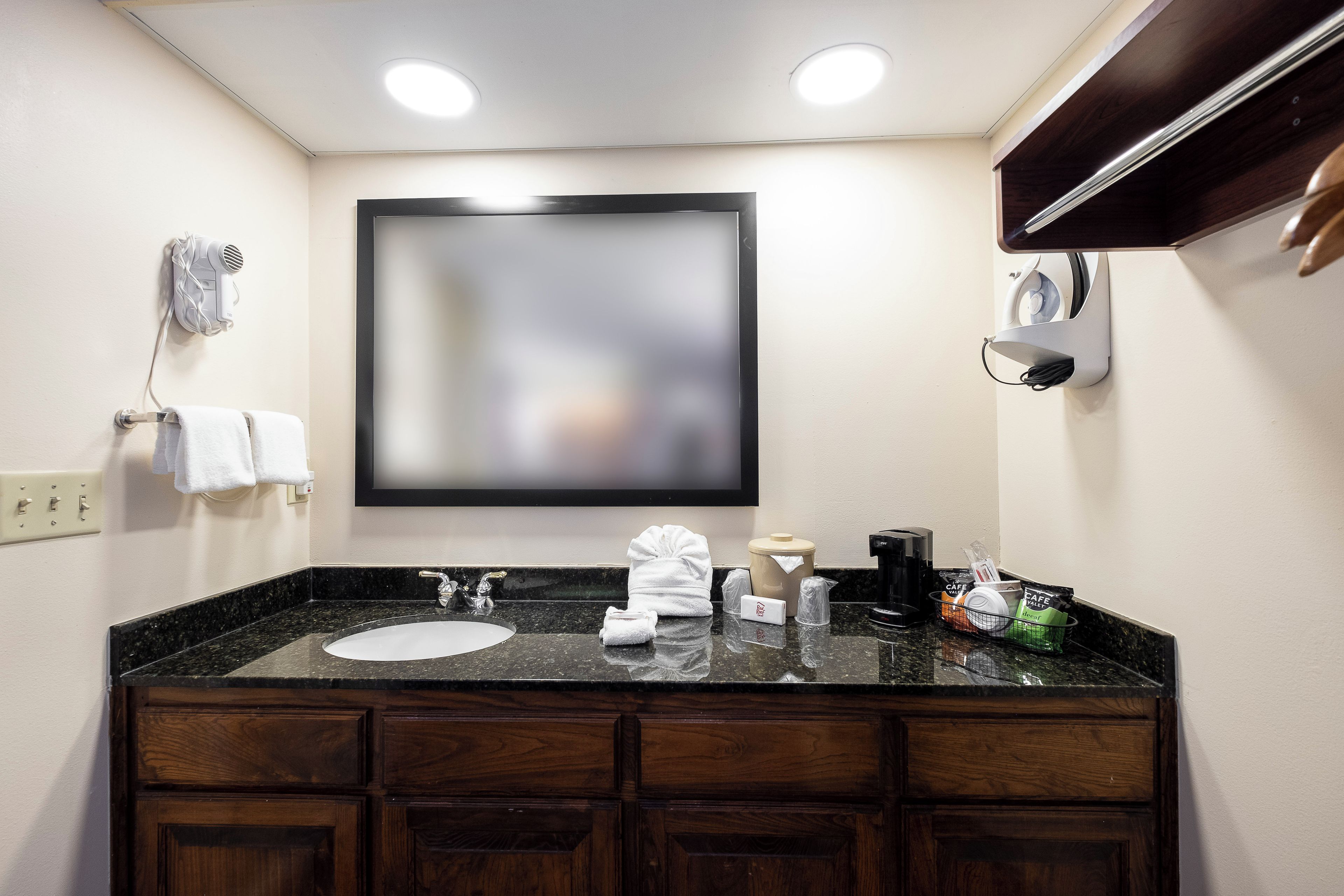 A hotel bathroom with a black granite countertop, a large mirror, a towel rack, a coffee maker, and toiletries.