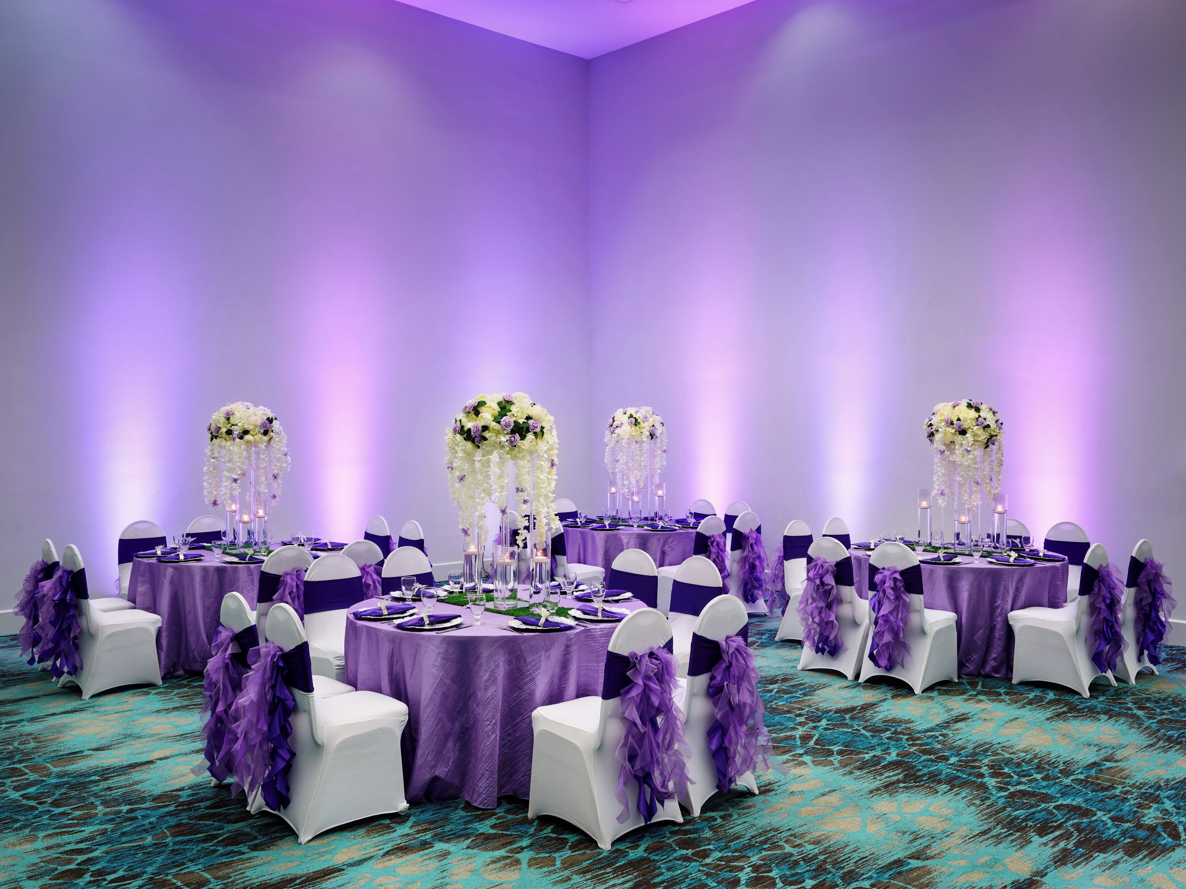 Ein Raum mit runden Tischen, violetten Tischdecken, weißen Stühlen mit violetten Bändern und floralen Tischdekorationen.