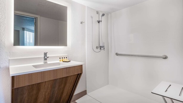 A modern bathroom with a white sink, wooden vanity, a large mirror, a shower with a handheld showerhead, and a grab bar.