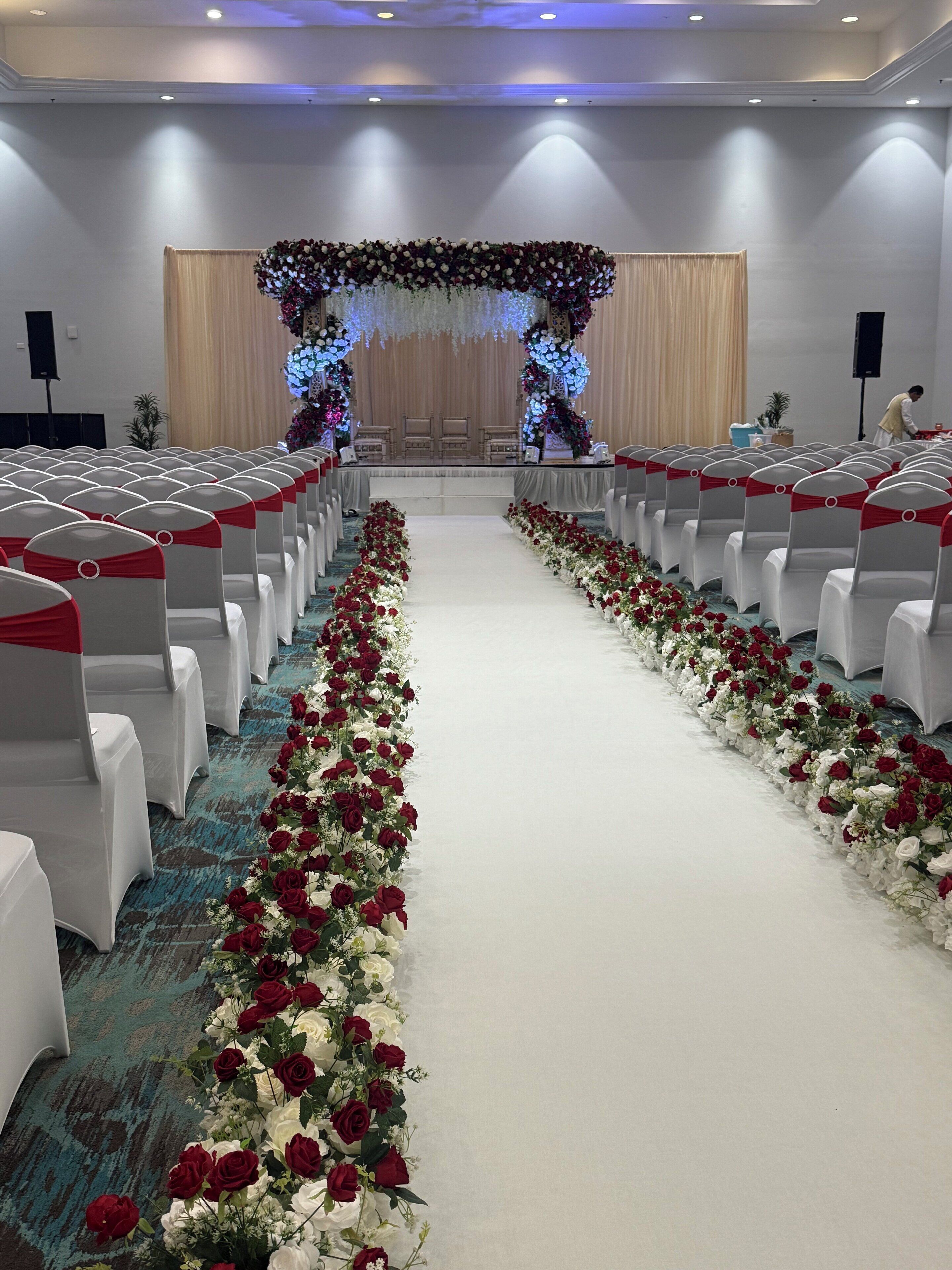 A hotel conference room with rows of white chairs, red sashes, and a floral aisle.
