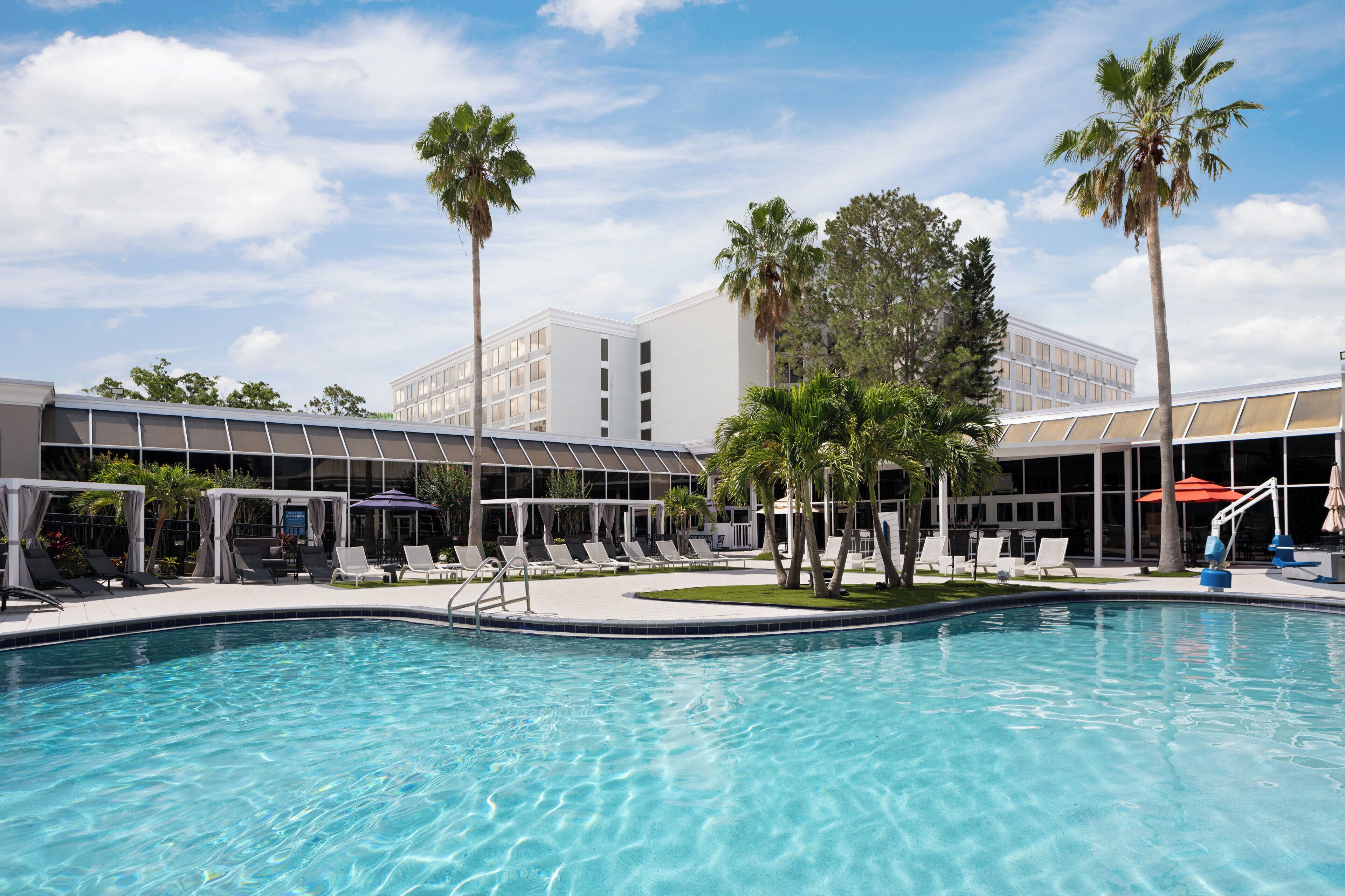 A swimming pool with lounge chairs and umbrellas, surrounded by palm trees and modern buildings.