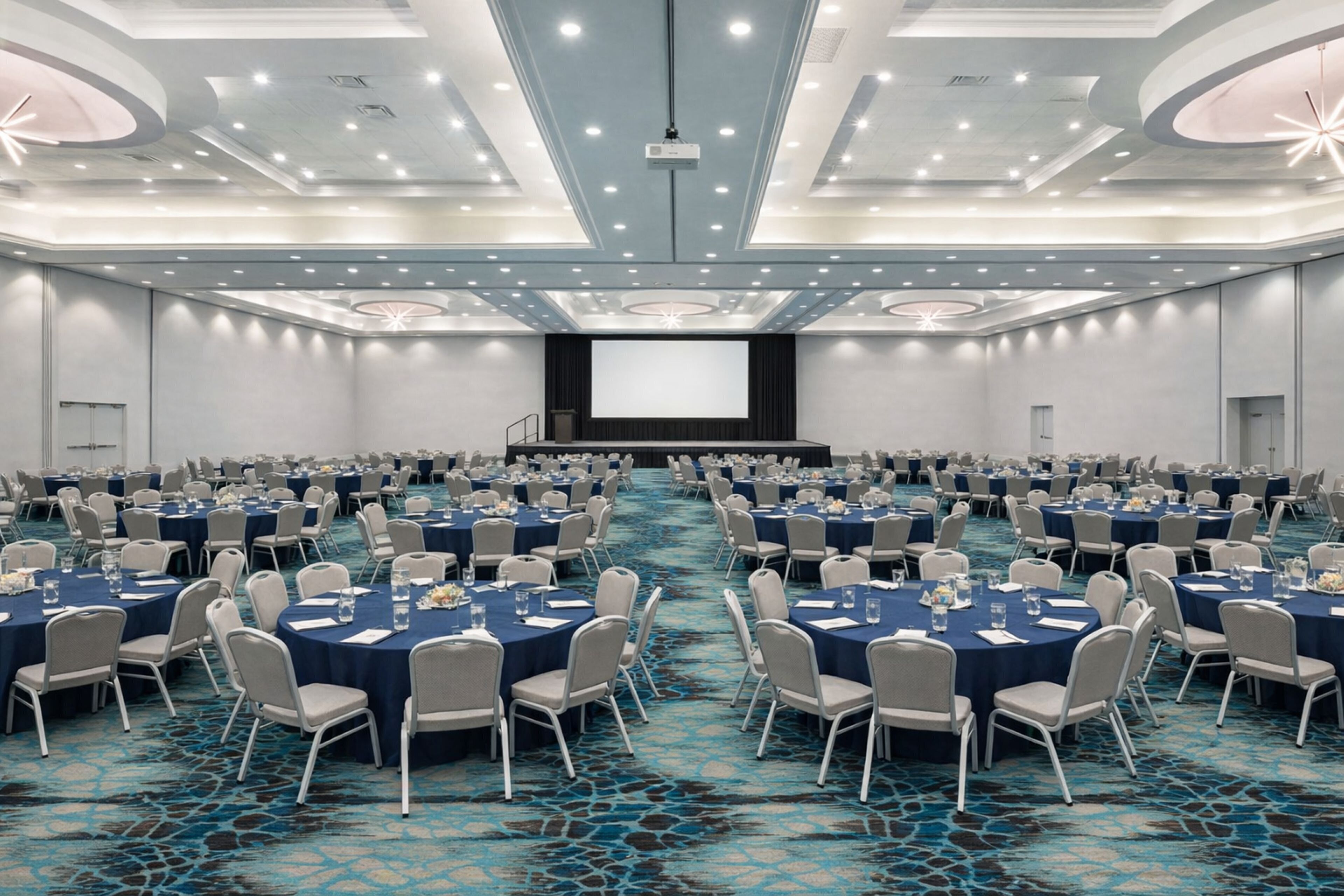 Une grande salle de conférence équipée de tables rondes et de chaises, d’une scène et d’un grand écran.