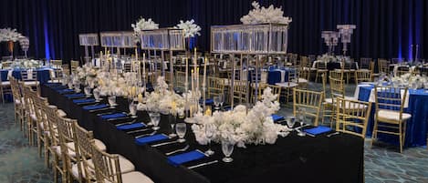 Un salón de banquetes con mesas largas, sillas doradas y manteles azules.