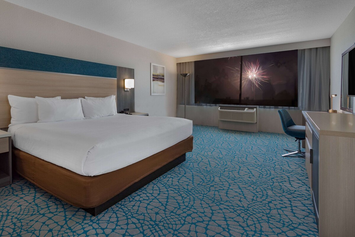A hotel room with a large bed, a desk, a chair, a large window with a view, and a wall-mounted air conditioner. - Holiday Inn Resort Kissimmee by the Parks by IHG (Kissimmee)