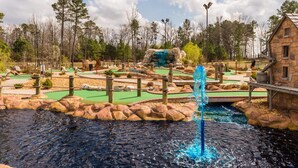 A miniature golf course with a water fountain, a stone building, and a forested area in the background.