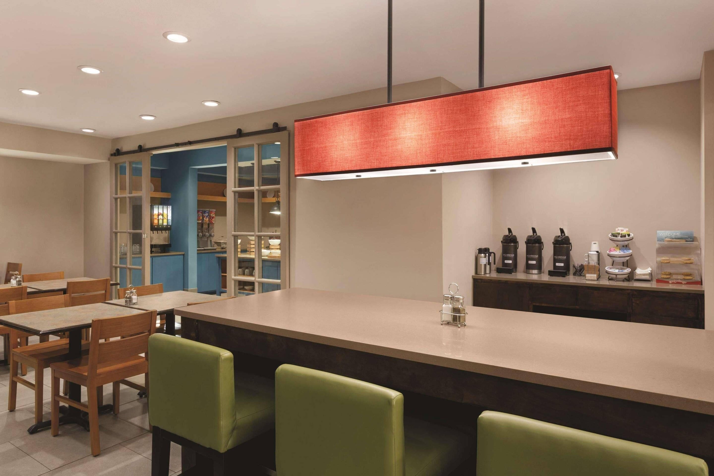 A modern dining area with a long table, green chairs, a coffee station, and a view of a blue room through glass doors.