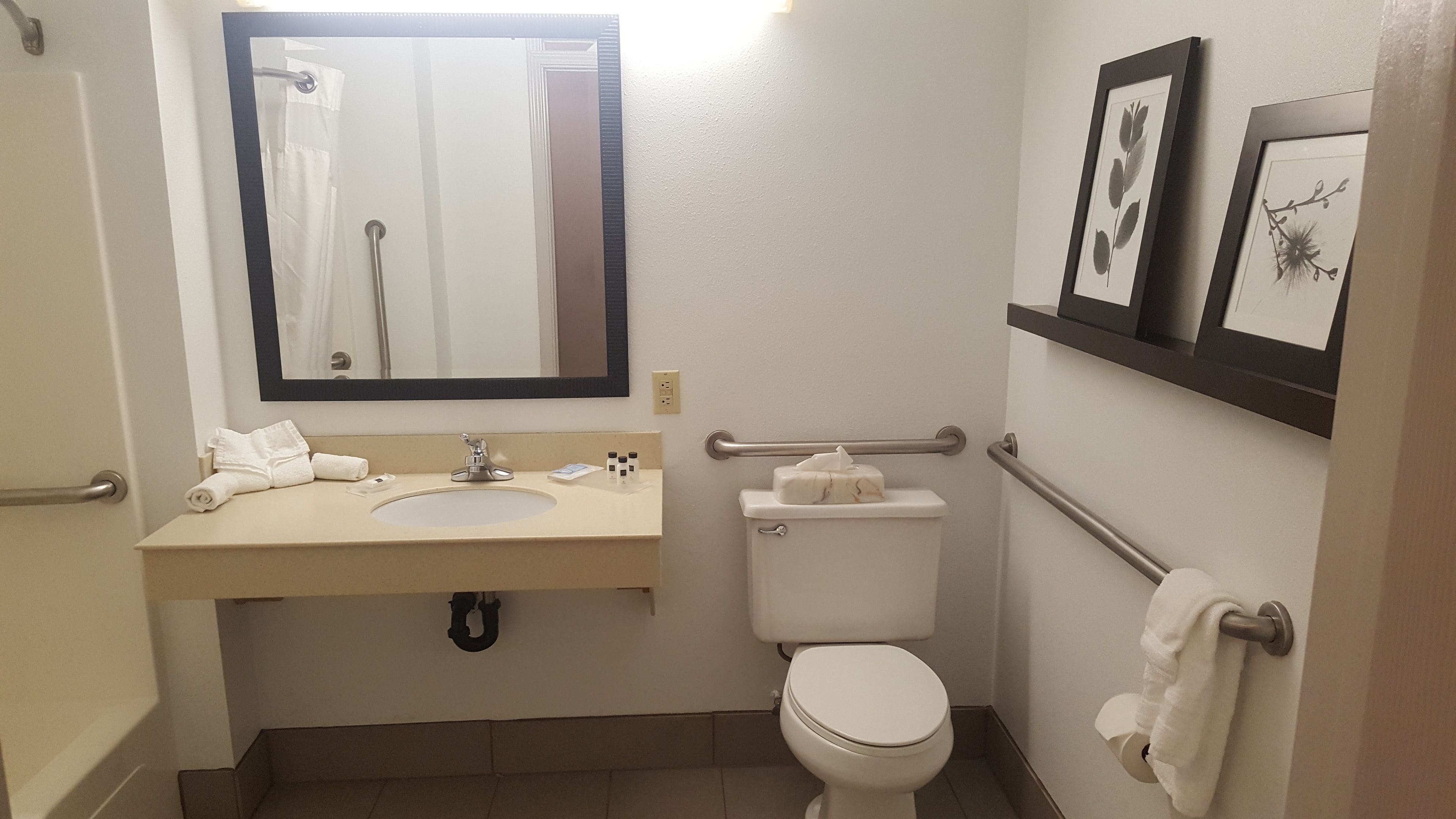 Un baño pequeño y limpio con lavamanos, espejo, inodoro y barras de apoyo.
