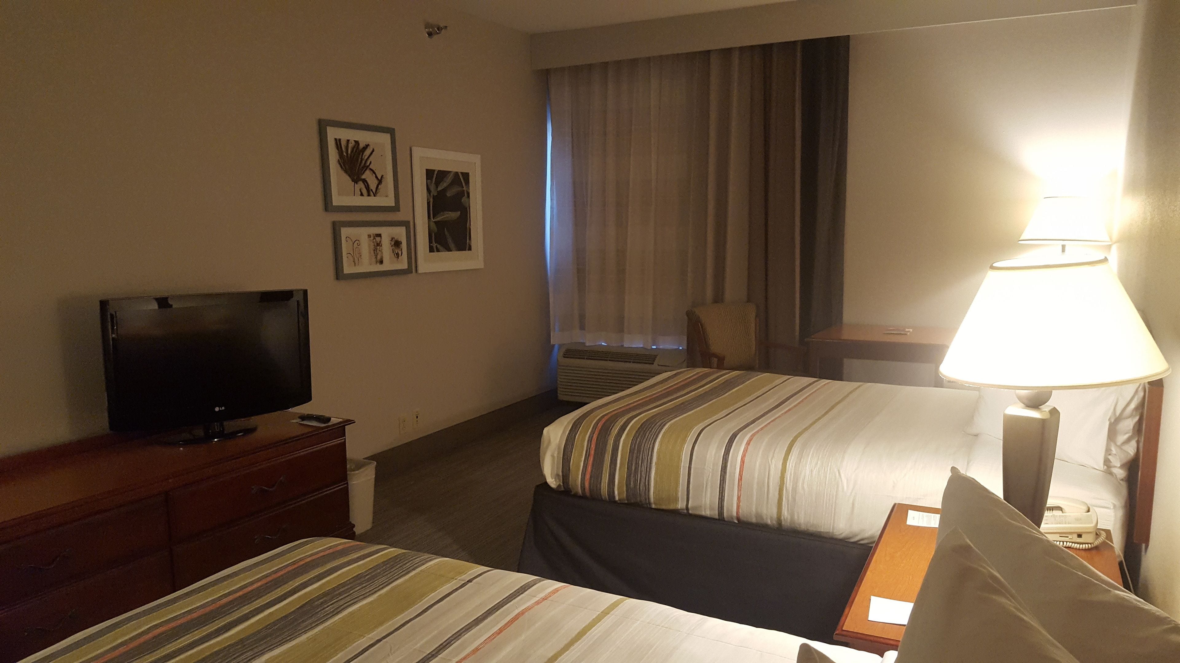 A hotel room with two beds, a TV on a dresser, a lamp, and framed pictures on the wall.