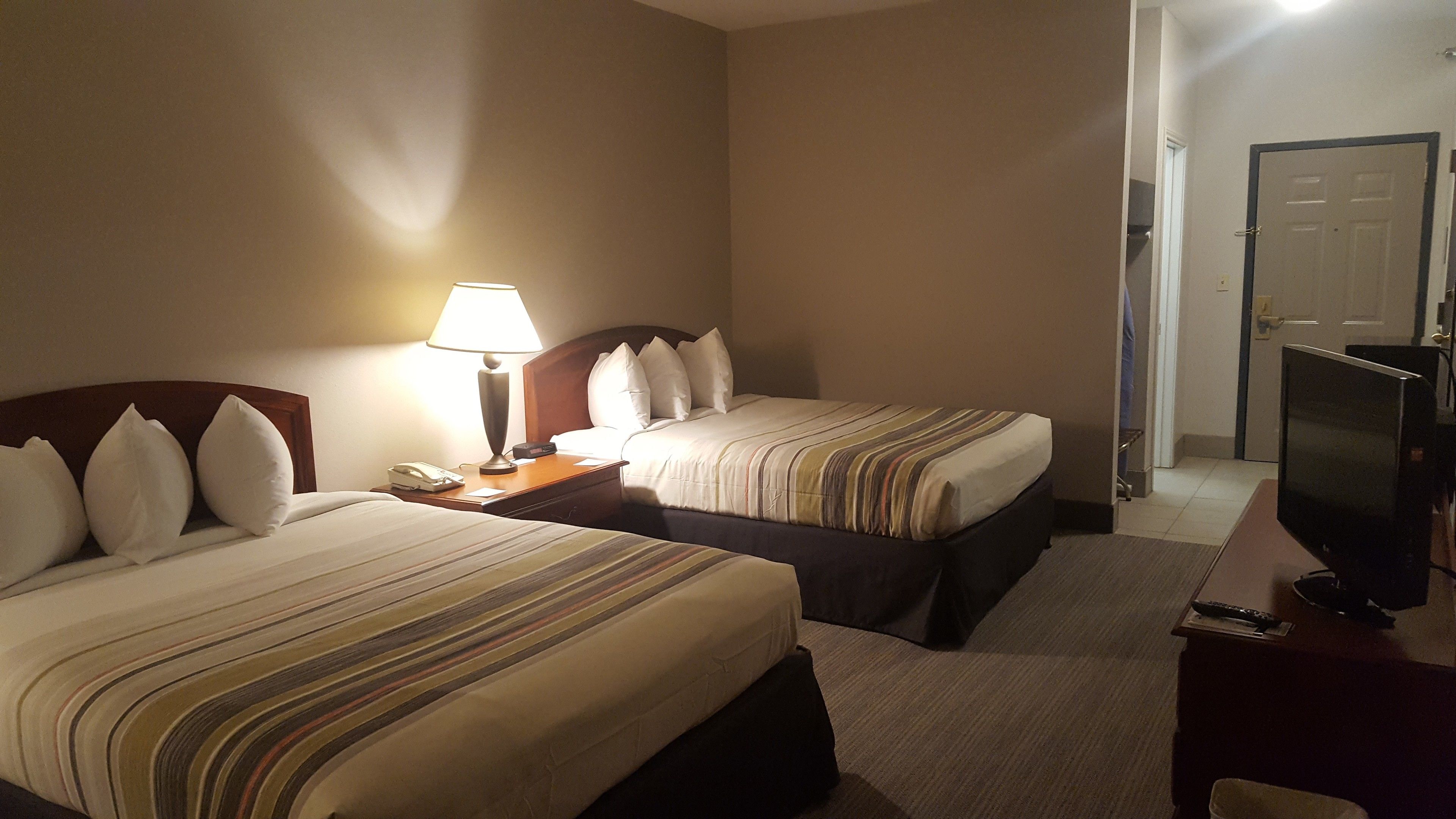 A hotel room with two beds, a desk, a lamp, a TV, and a door.