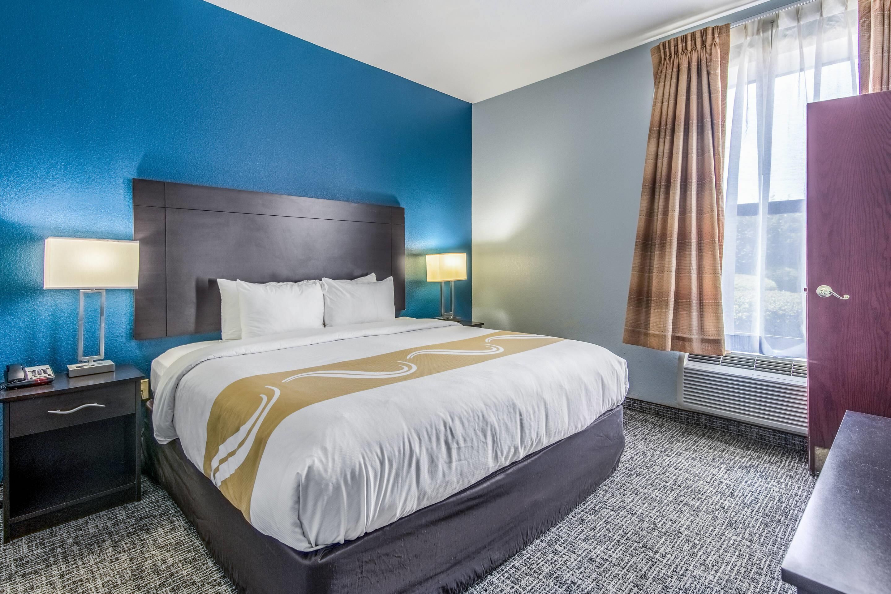 A hotel room with a large bed, two bedside tables with lamps, a window with curtains, and a door.