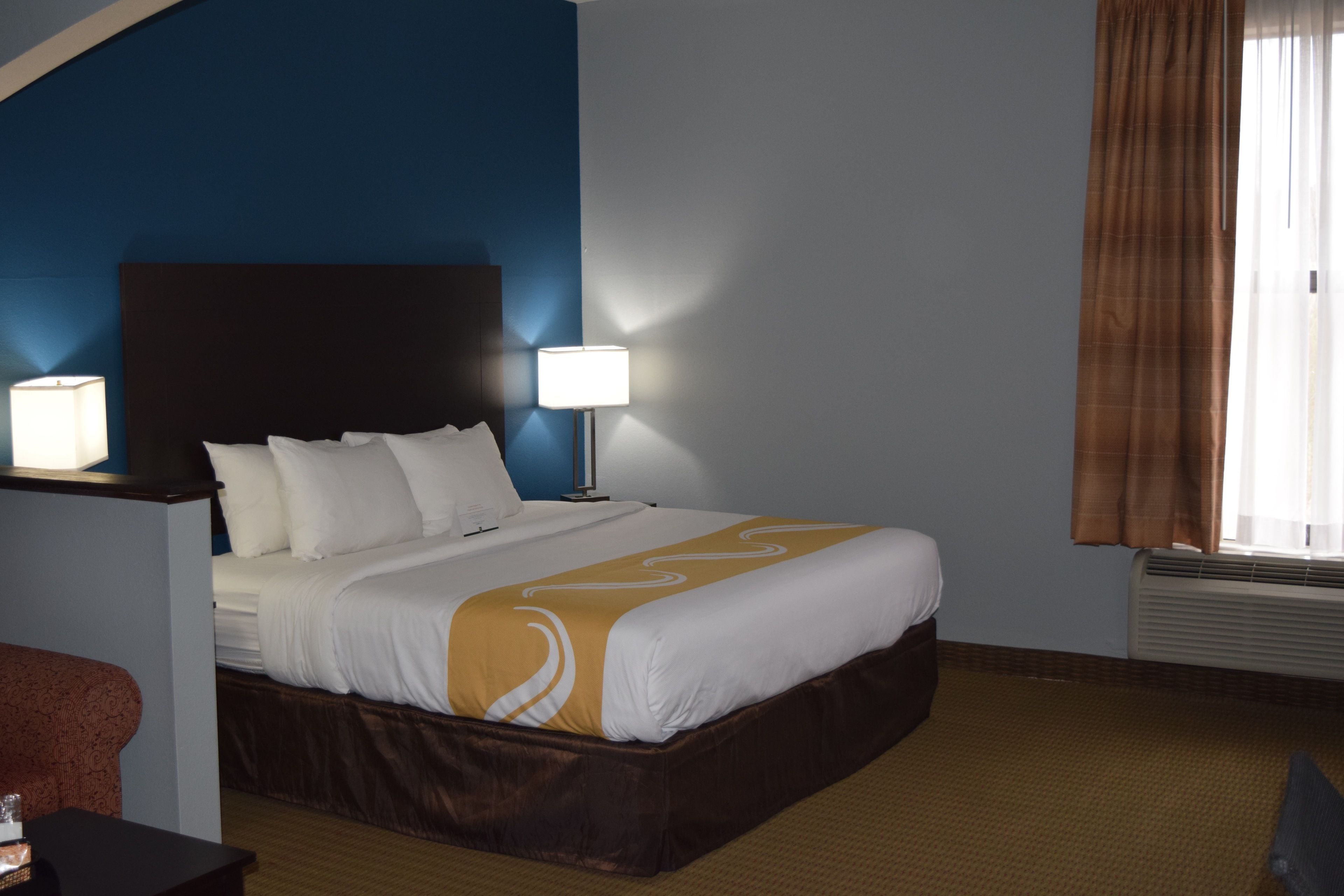 A hotel room with a bed, two bedside lamps, a brown headboard, a window with curtains, and a carpeted floor.