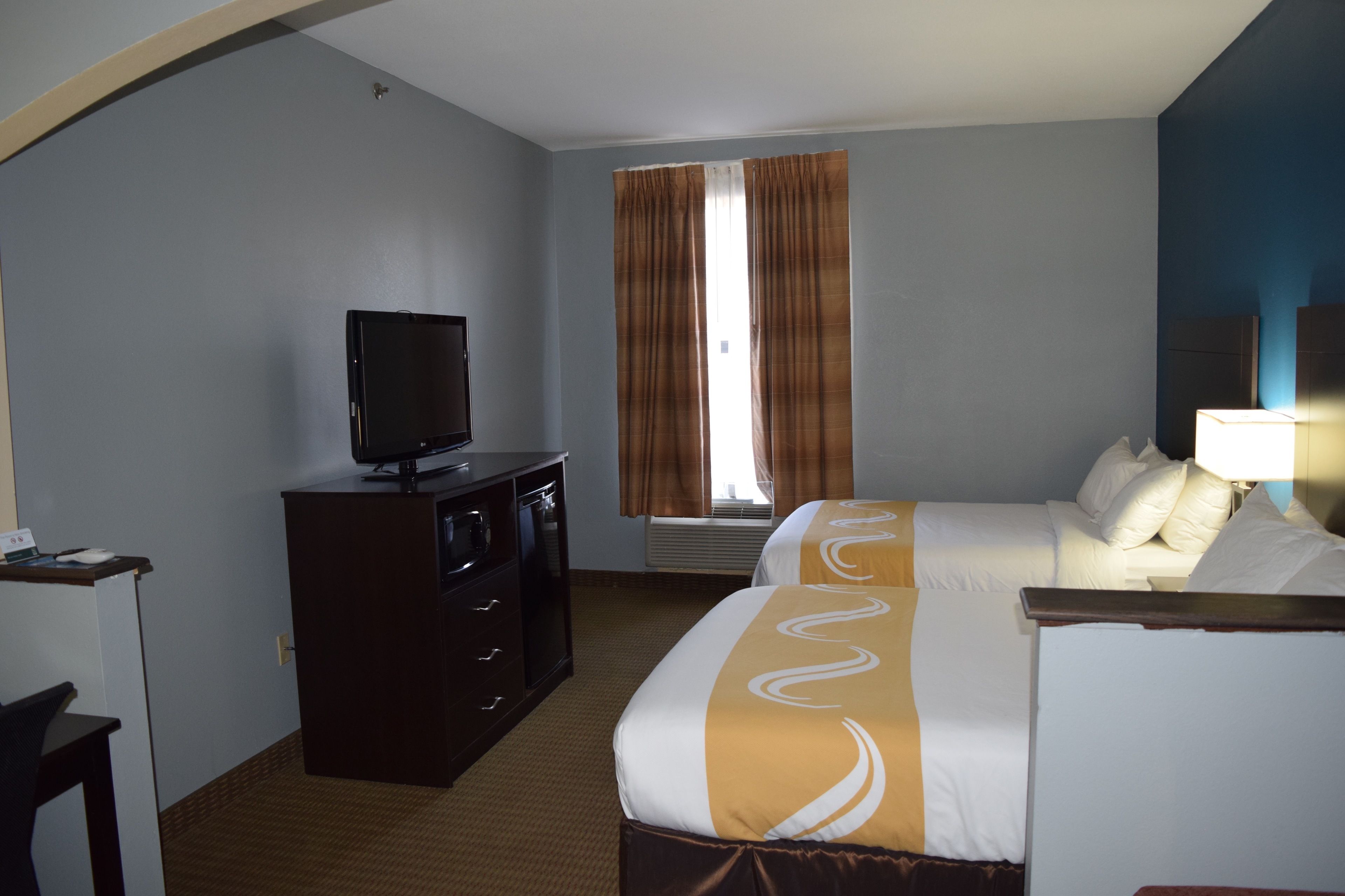 A hotel room with a bed, a TV on a dresser, a window with curtains, and a lamp on the bedside table.