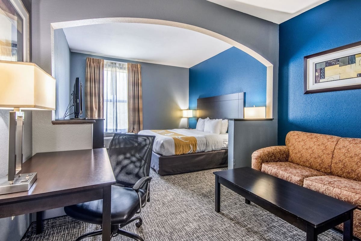 A hotel room with a bed, desk, chair, sofa, and a large window with curtains.