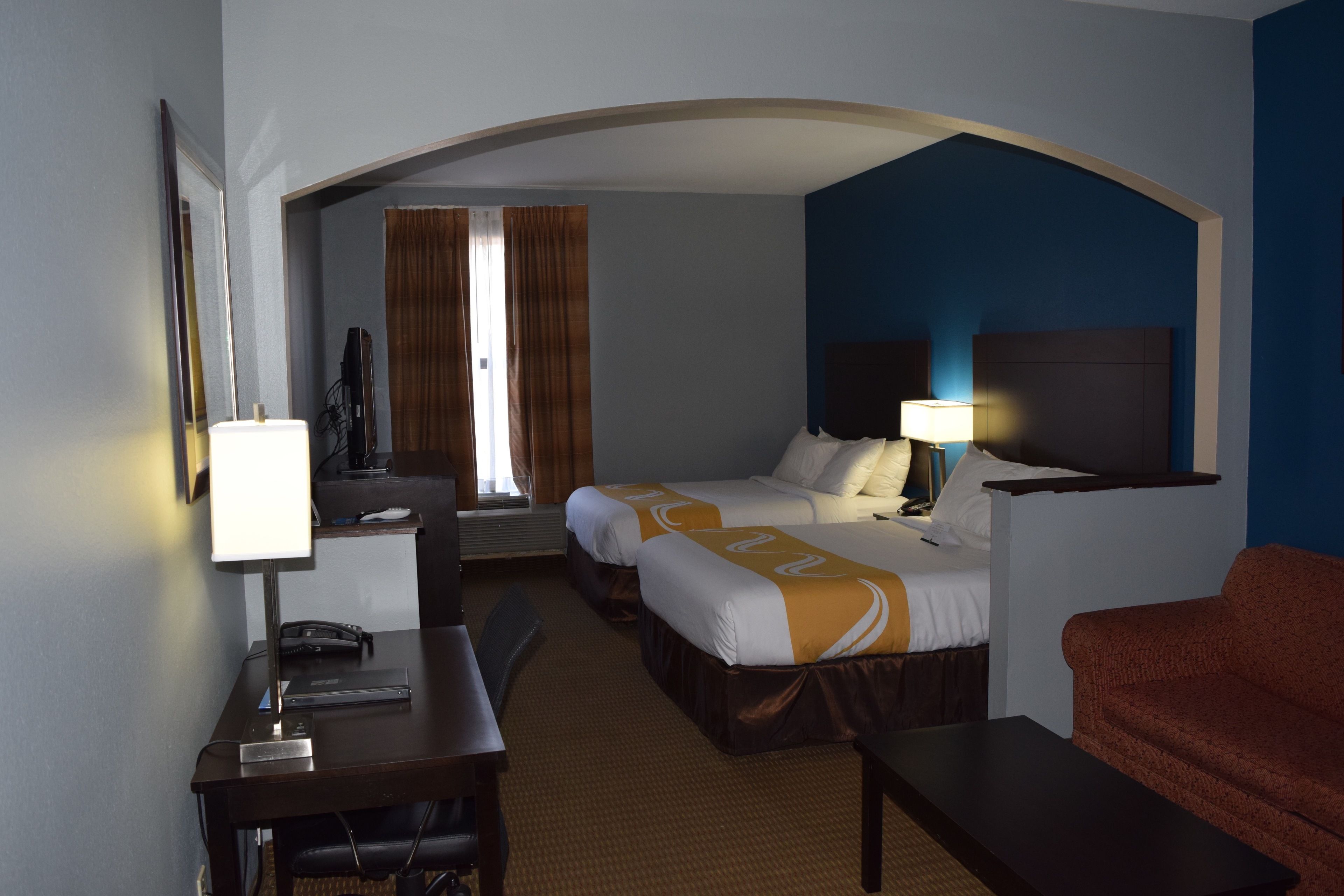 Quarto de hotel com duas camas, uma escrivaninha, uma cadeira, uma televisão e um sofá.