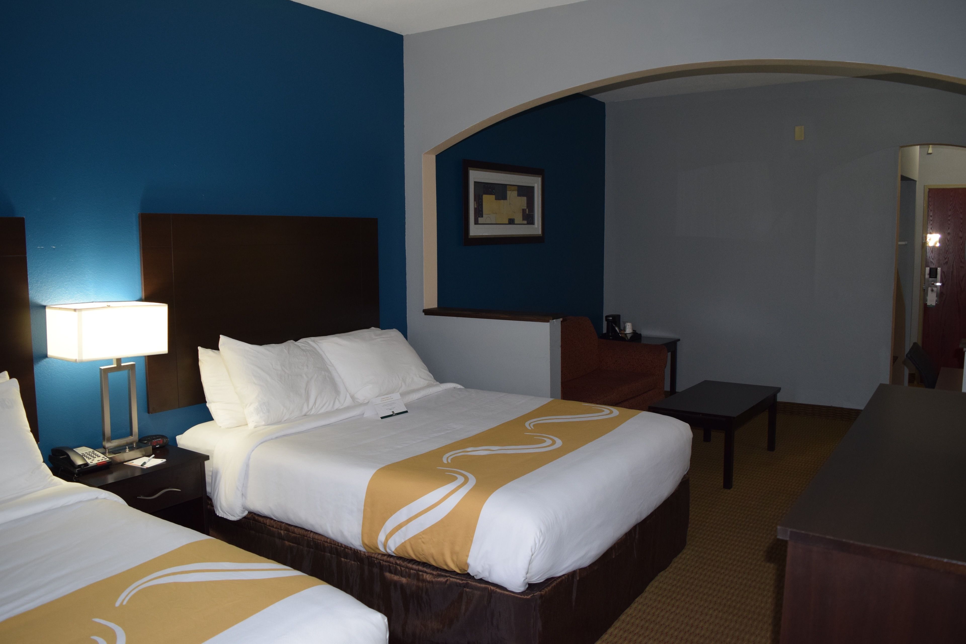A hotel room with two beds, a desk, a chair, and a nightstand with a lamp.