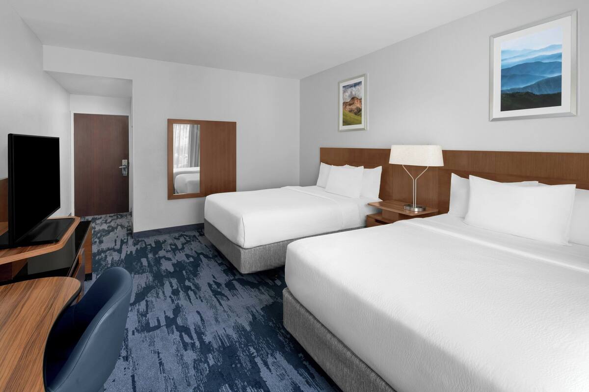 A hotel room with two beds, a desk with a chair, a TV, and a large window.