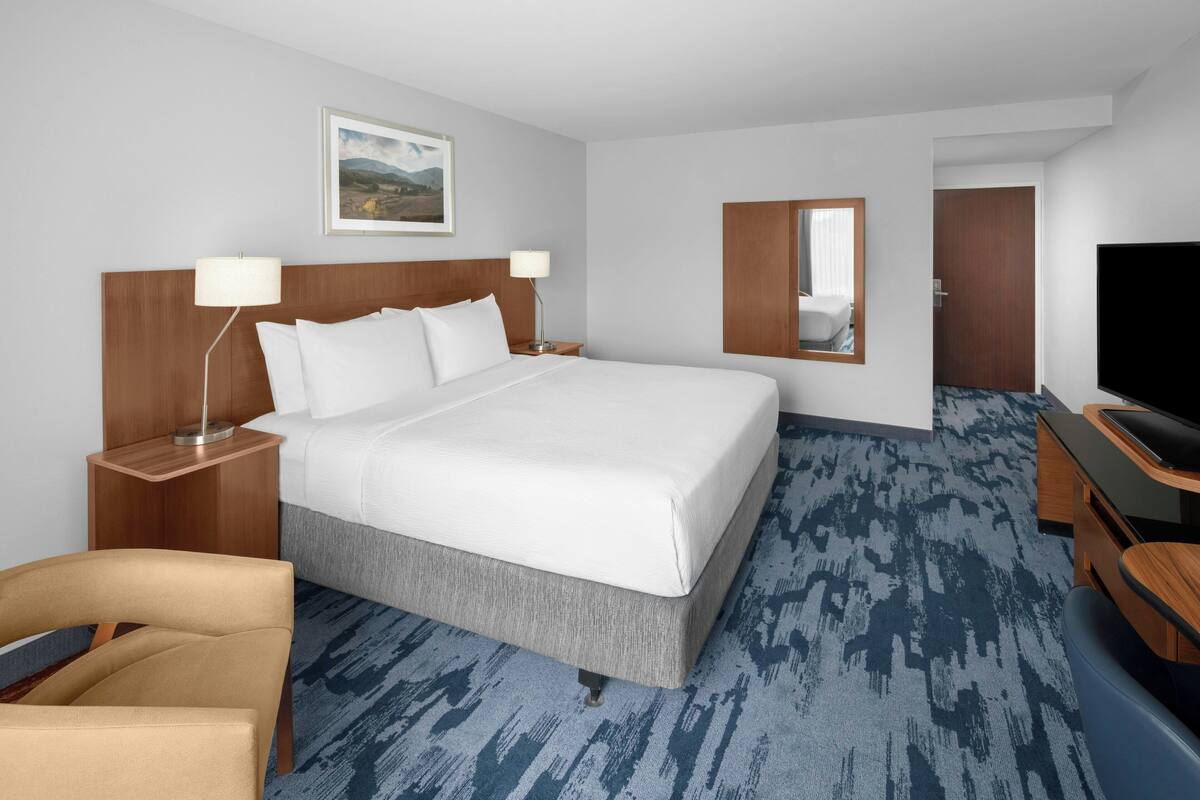 A hotel room with a large bed, two bedside tables with lamps, a TV, a chair, and a carpeted floor.